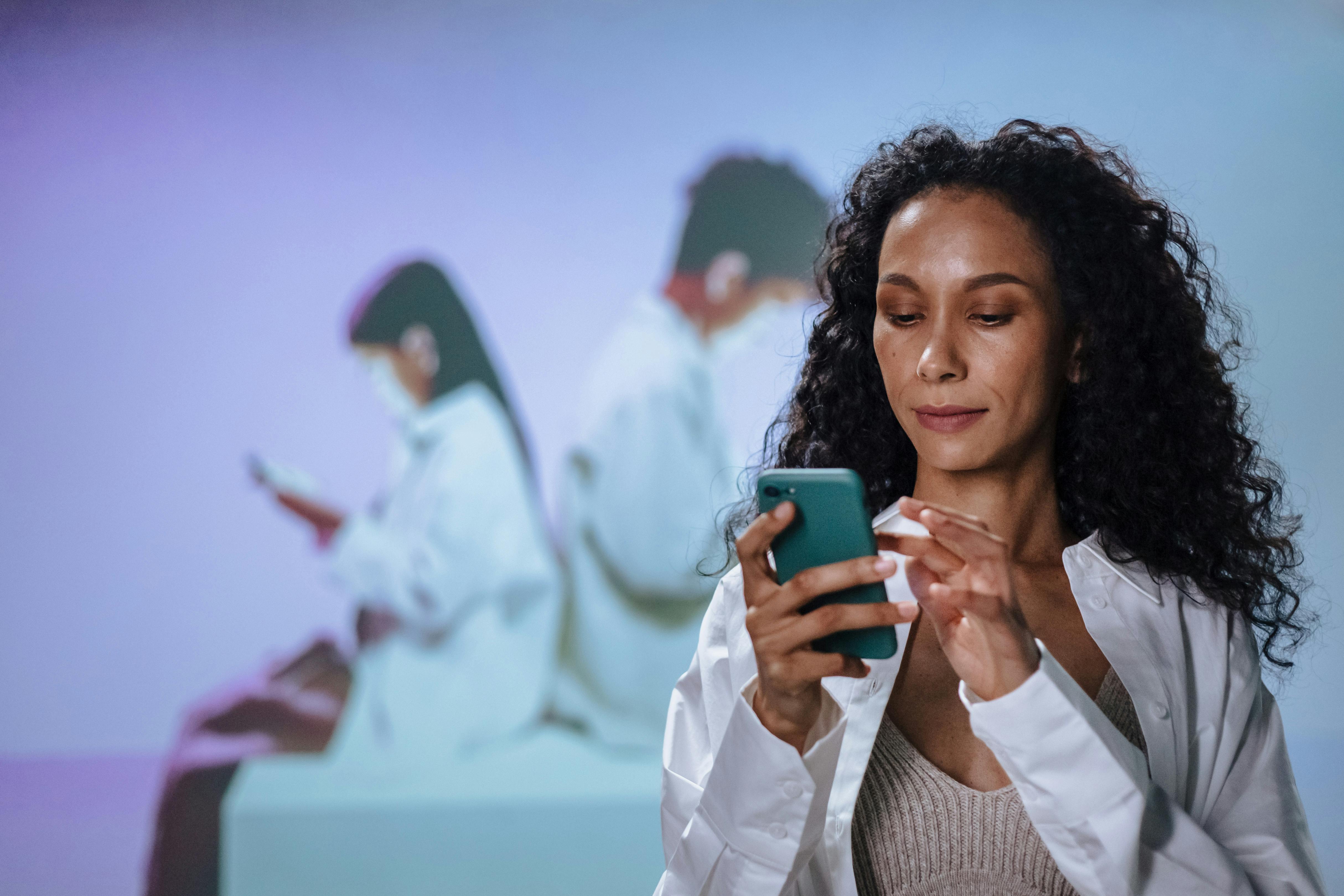 A woman (doctor) with curly hair looks at her smartphone in the foreground, with a blurred projection of people using mobile devices in the background.
