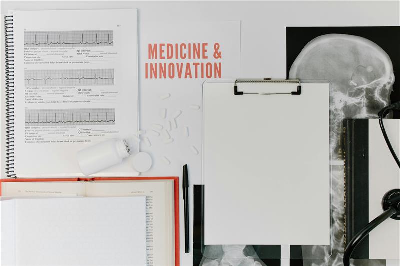 Flat lay of medical and research materials including an open book, printed ECG charts, scattered pills, a blank clipboard, a pen, a stethoscope, and a skull X-ray, with the words ‘Medicine & Innovation’ displayed in the center.